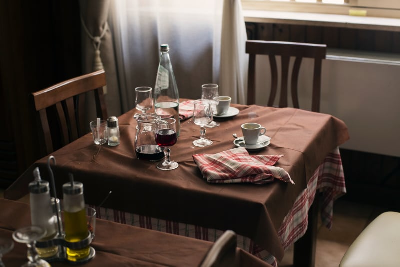 A finished meal on a brown-clothed restaurant table by a window: empty wine glasses, a water bottle, espresso cups and a red checked napkin.